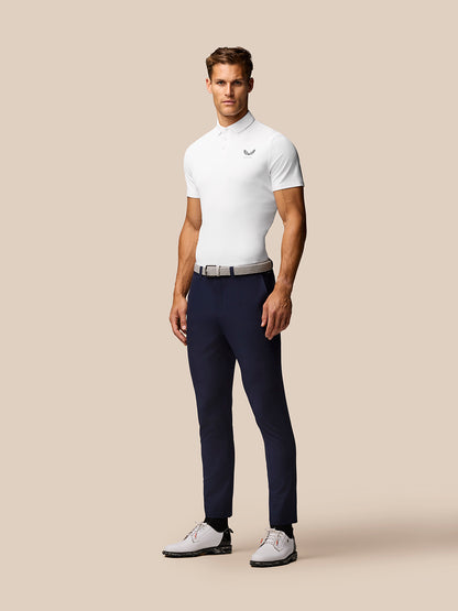 Male model wearing a white golf polo with Castore logo, navy trousers, belt, and white golf shoes against a beige background.