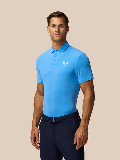 Male model wearing cool blue Golf Club Essential Polo with Castore logo on chest against a beige background.