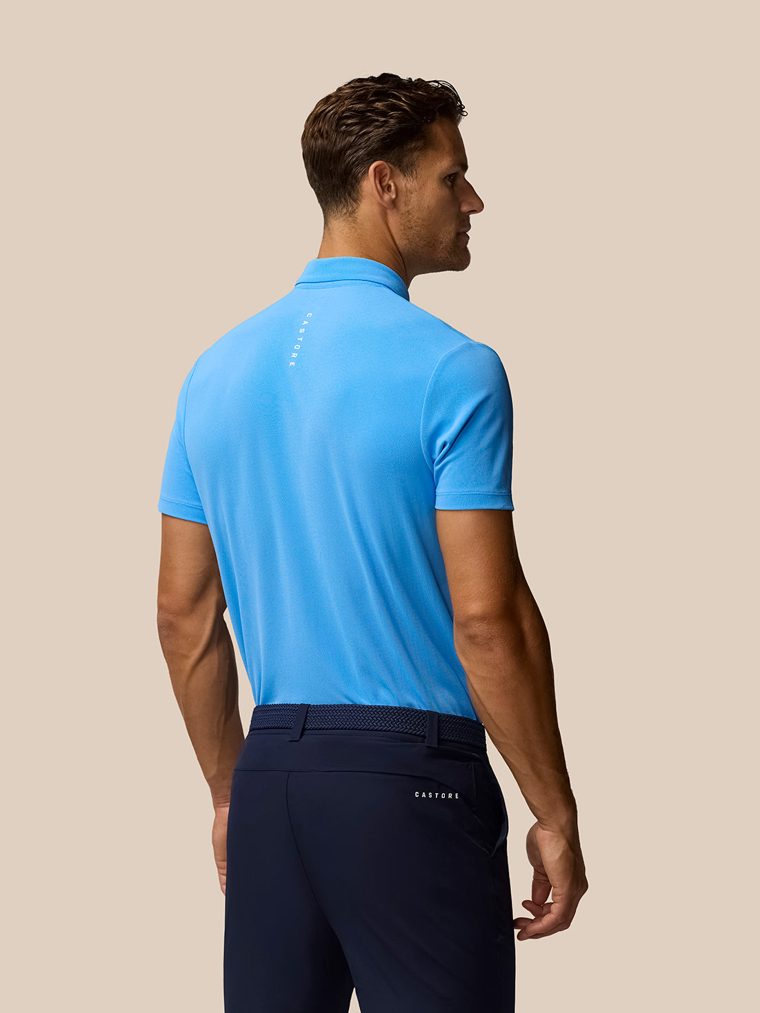 Back view of a man in a light blue Castore Golf Club Essential Polo paired with navy shorts