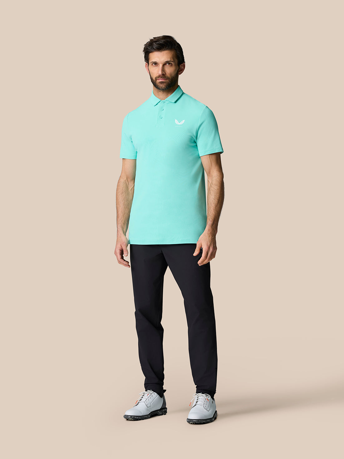 Man wearing light aqua Golf Club Essential Polo paired with dark pants and white golf shoes against a neutral studio backdrop.