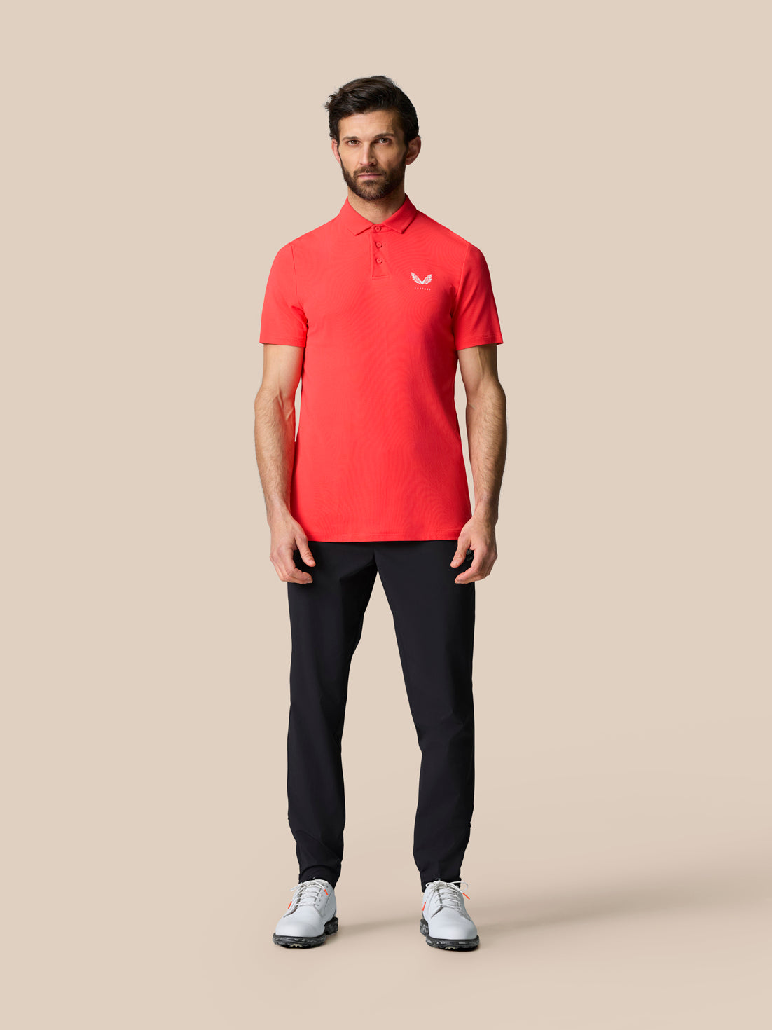 Male model wearing red Golf Club Essential Polo with black trousers and white sneakers, Castore logo on the chest