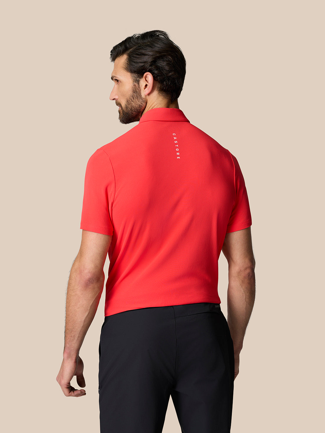 Back view of a man wearing a red Golf Club Essential Polo with vertical Castore branding on the upper back.