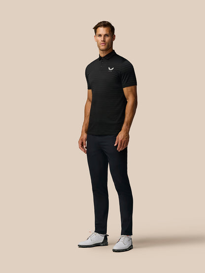 Full-length shot of a man in a black Pinnacle Pique Stripe Polo with striped knit, Castore logo on chest, paired with dark trousers and white golf shoes.