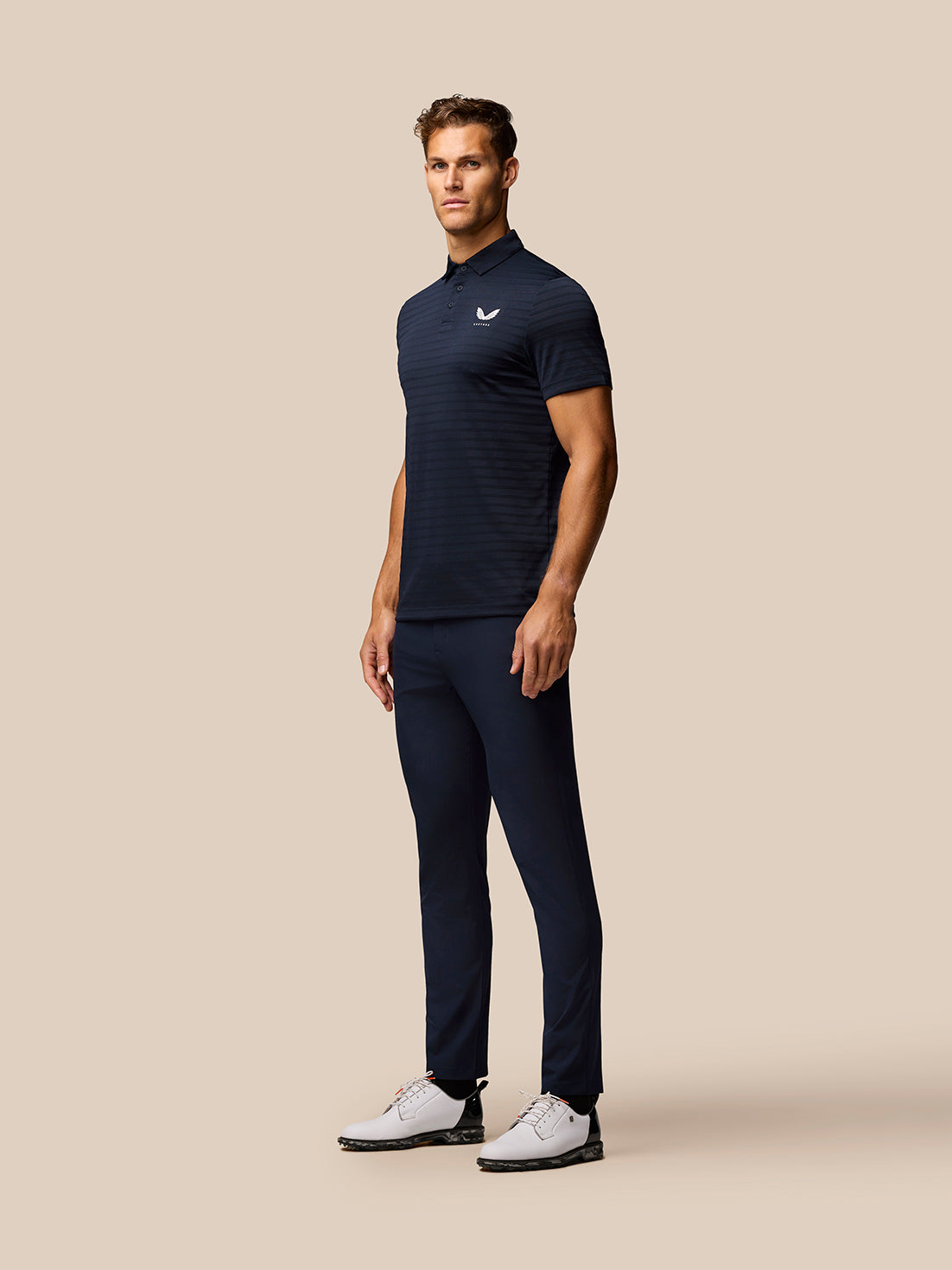 Male model in navy Pinnacle Pique Stripe Polo, short sleeves, three quarter view against beige backdrop.