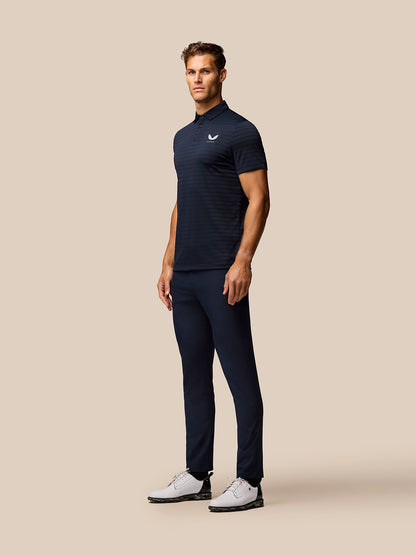 Male model in navy Pinnacle Pique Stripe Polo, short sleeves, three quarter view against beige backdrop.