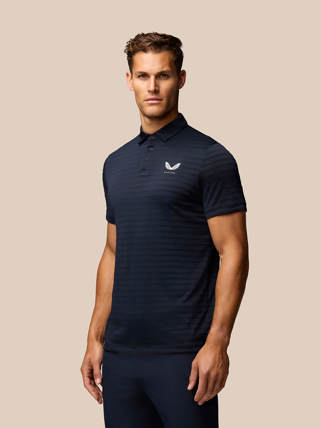 Male model wearing navy Pinnacle Pique Stripe Polo with stripe pattern and white logo on chest against a beige backdrop.
