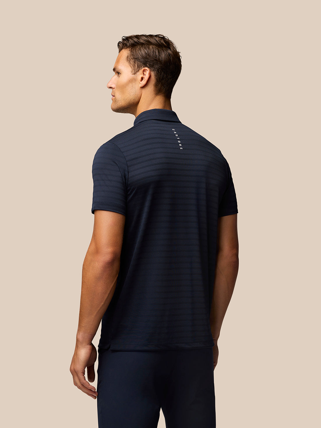 Back view of a man wearing navy Pinnacle Pique Stripe Polo with subtle stripes and Castore branding along the upper back.