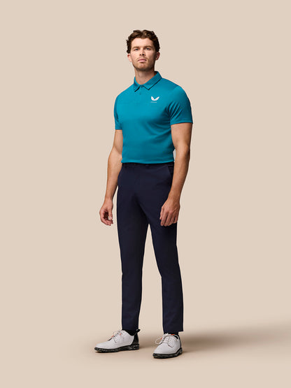 Full-body shot of a man wearing dark aqua Pinnacle golf polo with navy pants against a beige backdrop.