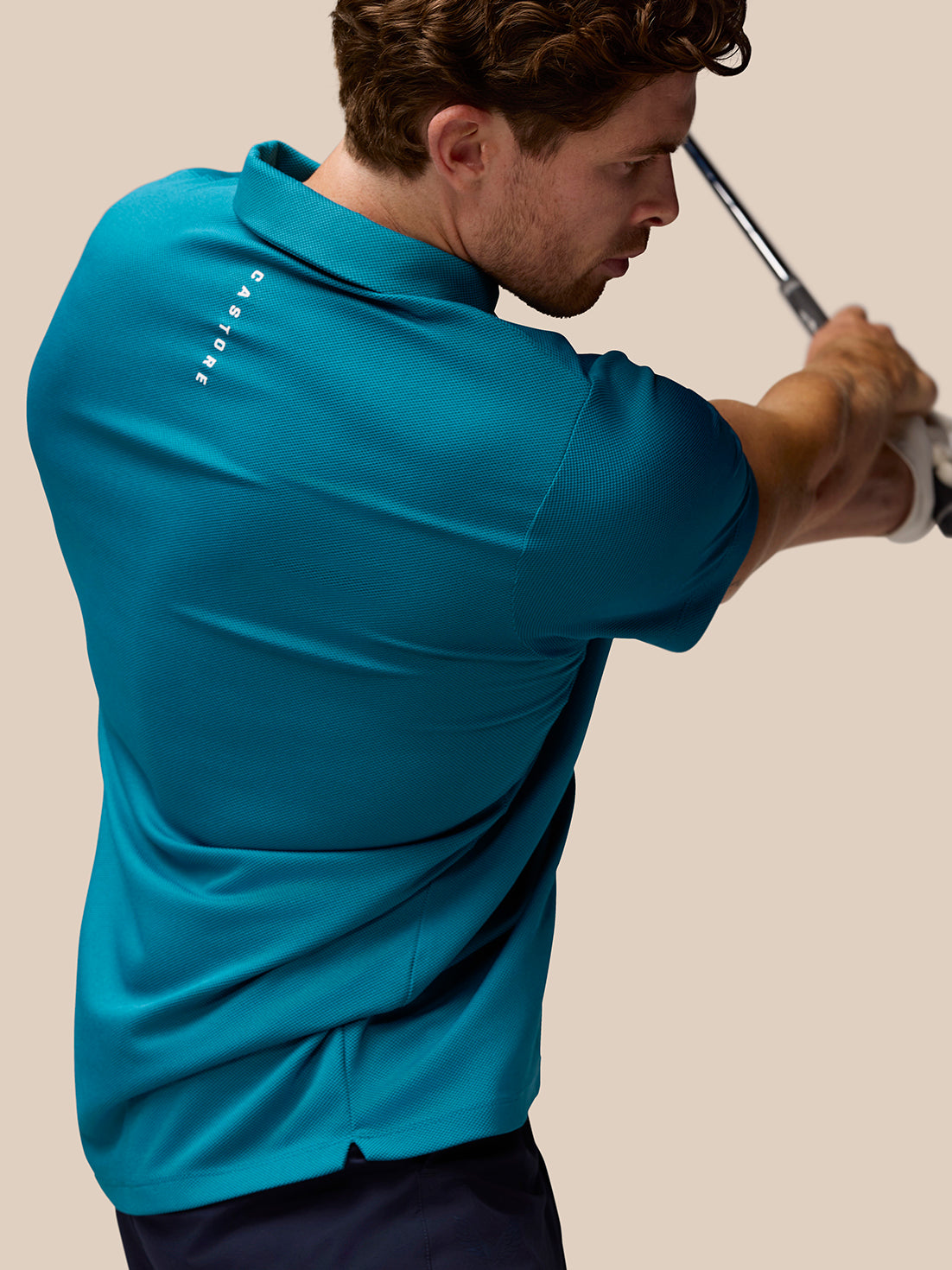 Back view of a man in dark aqua Pinnacle golf polo mid swing with a golf club.