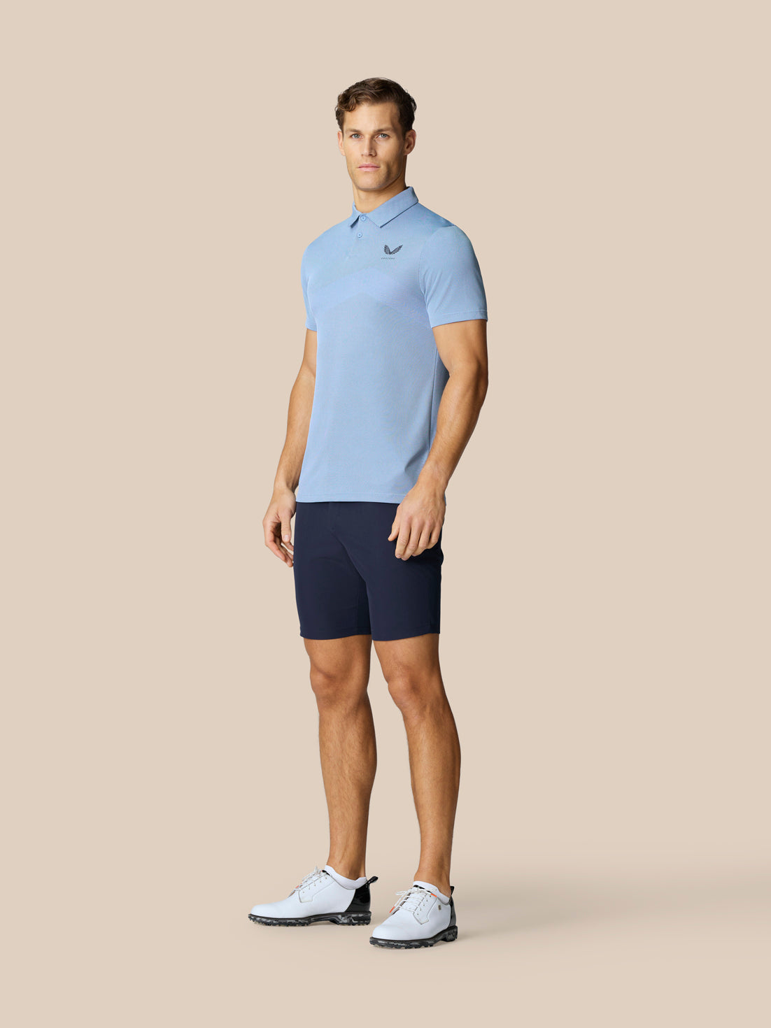 Male model in sky blue polo with navy shorts and white golf shoes, standing in a studio against a beige backdrop.
