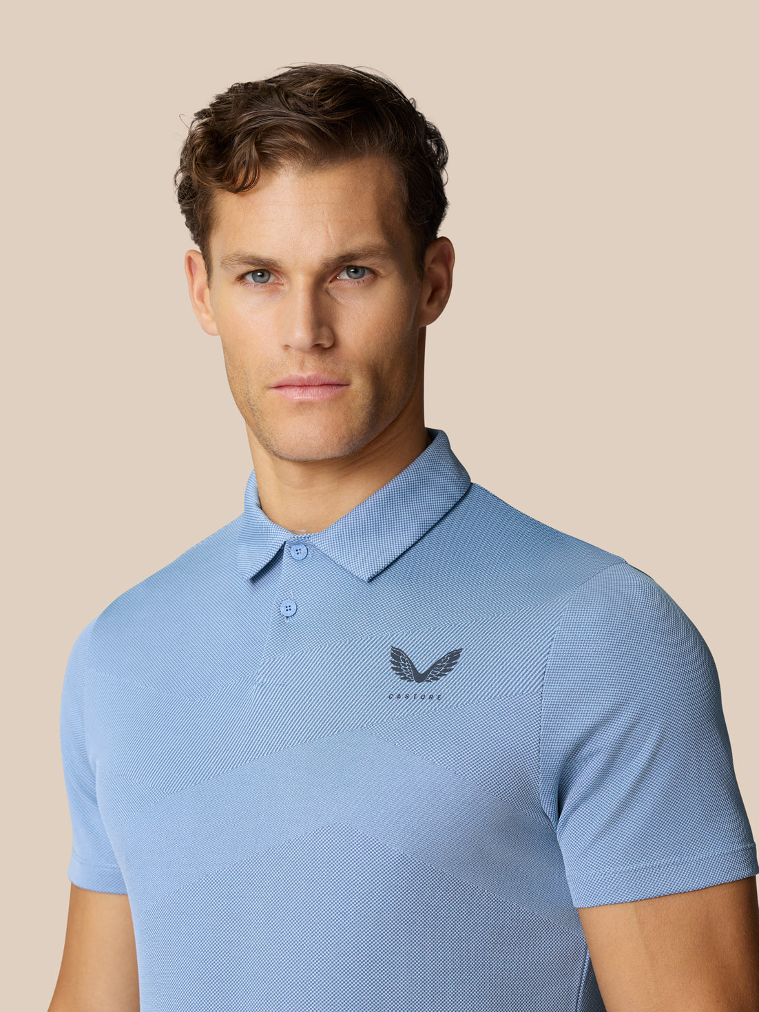 Male model wearing sky blue polo with chevron knit texture and chest logo, posed against a beige background.