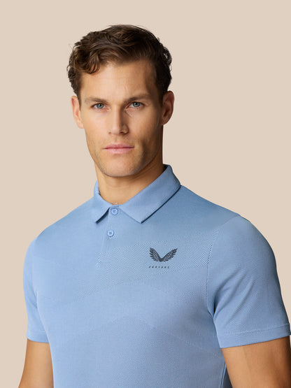 Male model wearing sky blue polo with chevron knit texture and chest logo, posed against a beige background.