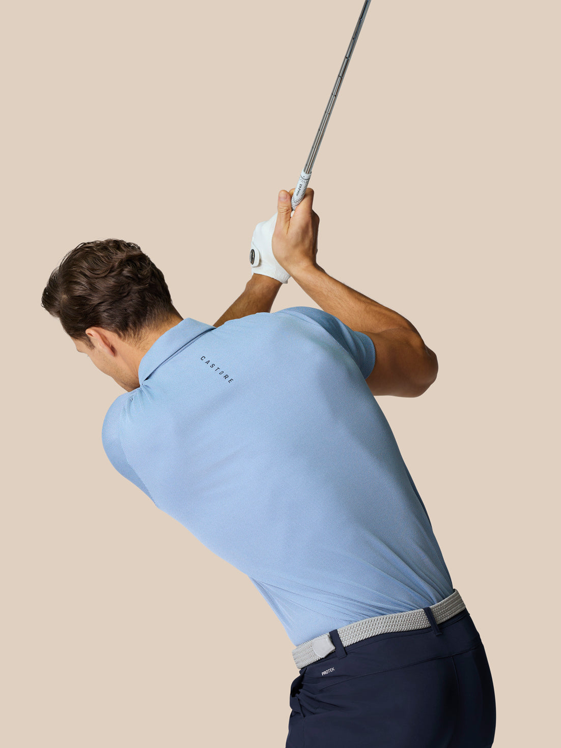 Golfer in sky blue polo mid swing with a raised club, showcasing athletic fit and back branding.