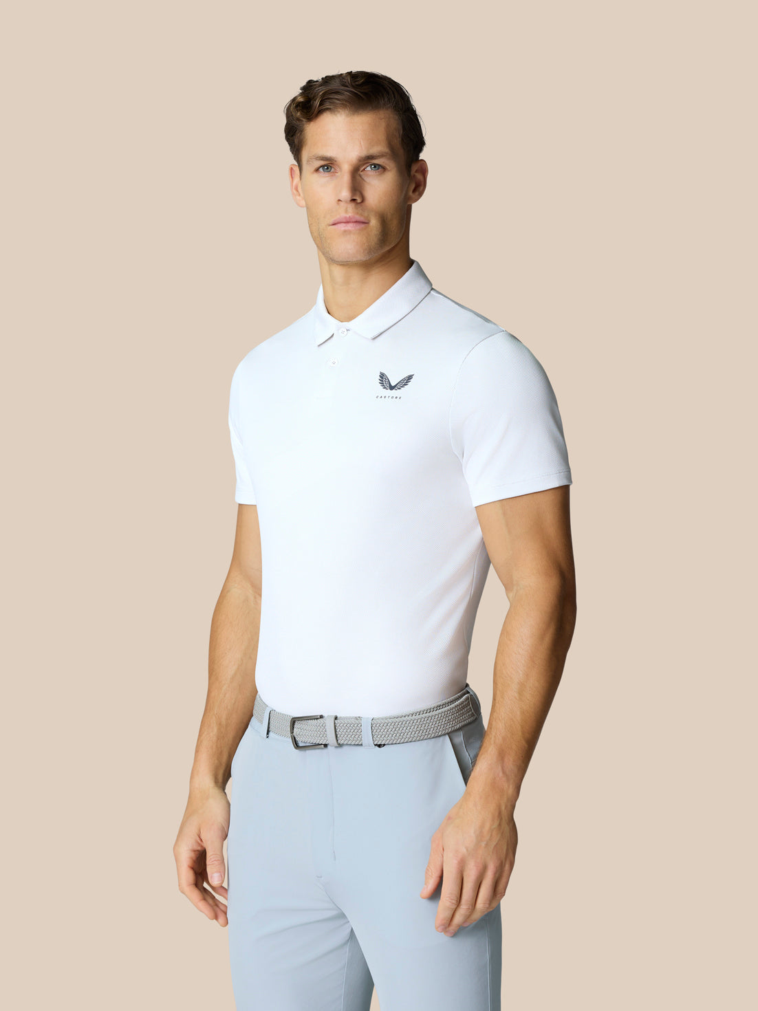 Male model wearing white Golf Pinnacle Polo with Castore logo on chest and light blue pants against a beige backdrop