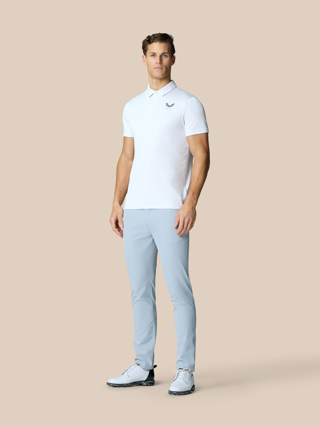 Male model wearing white Golf Pinnacle Polo with light blue pants on a beige background