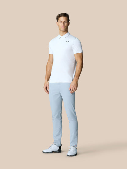 Male model wearing white Golf Pinnacle Polo with light blue pants on a beige background