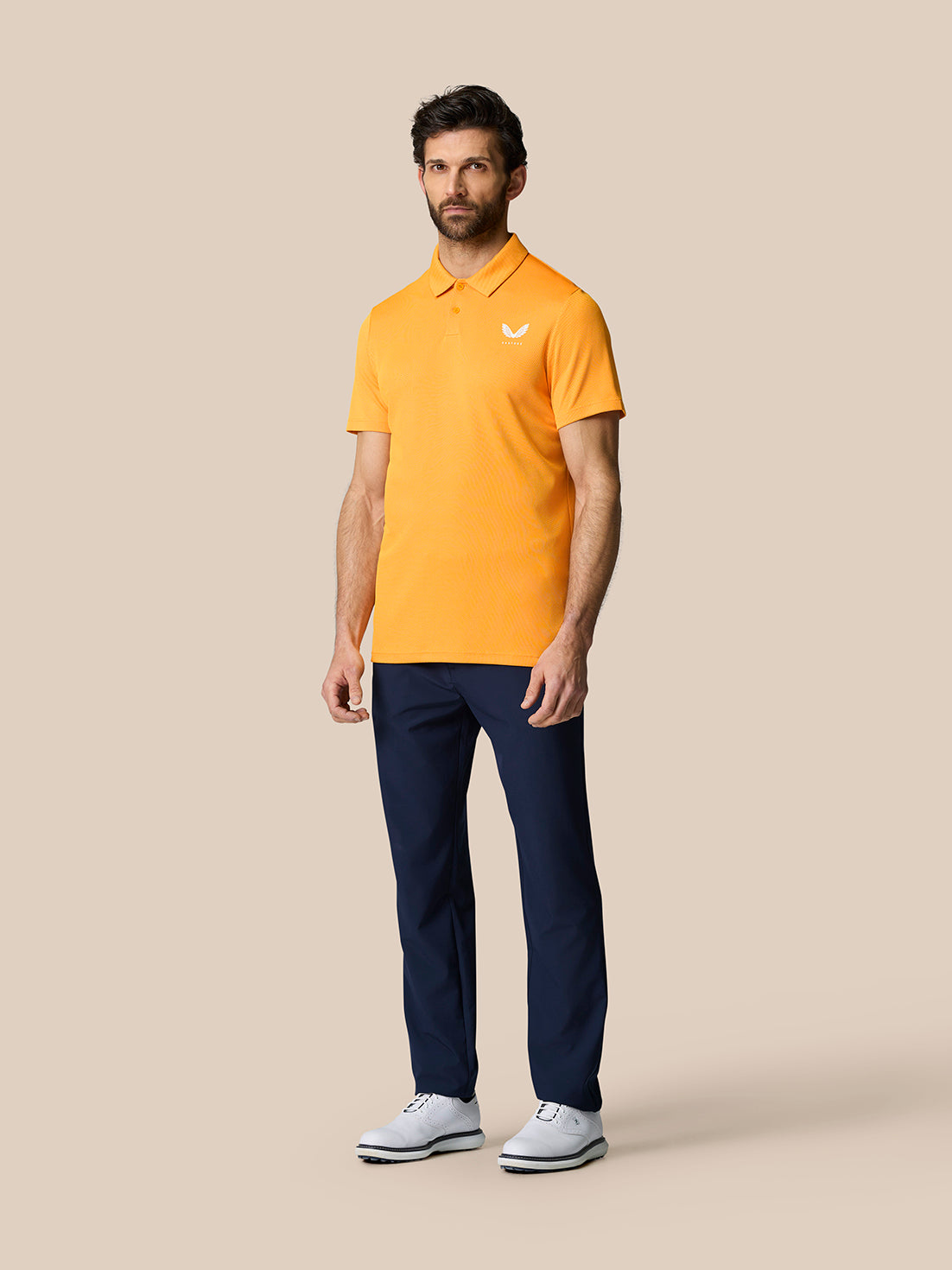 Male model wearing orange Golf Pinnacle Polo Mango with navy pants and white shoes.