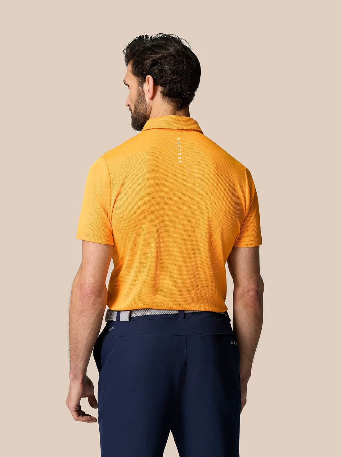 Rear view of male model in bright orange Golf Pinnacle Polo showing back Castore branding