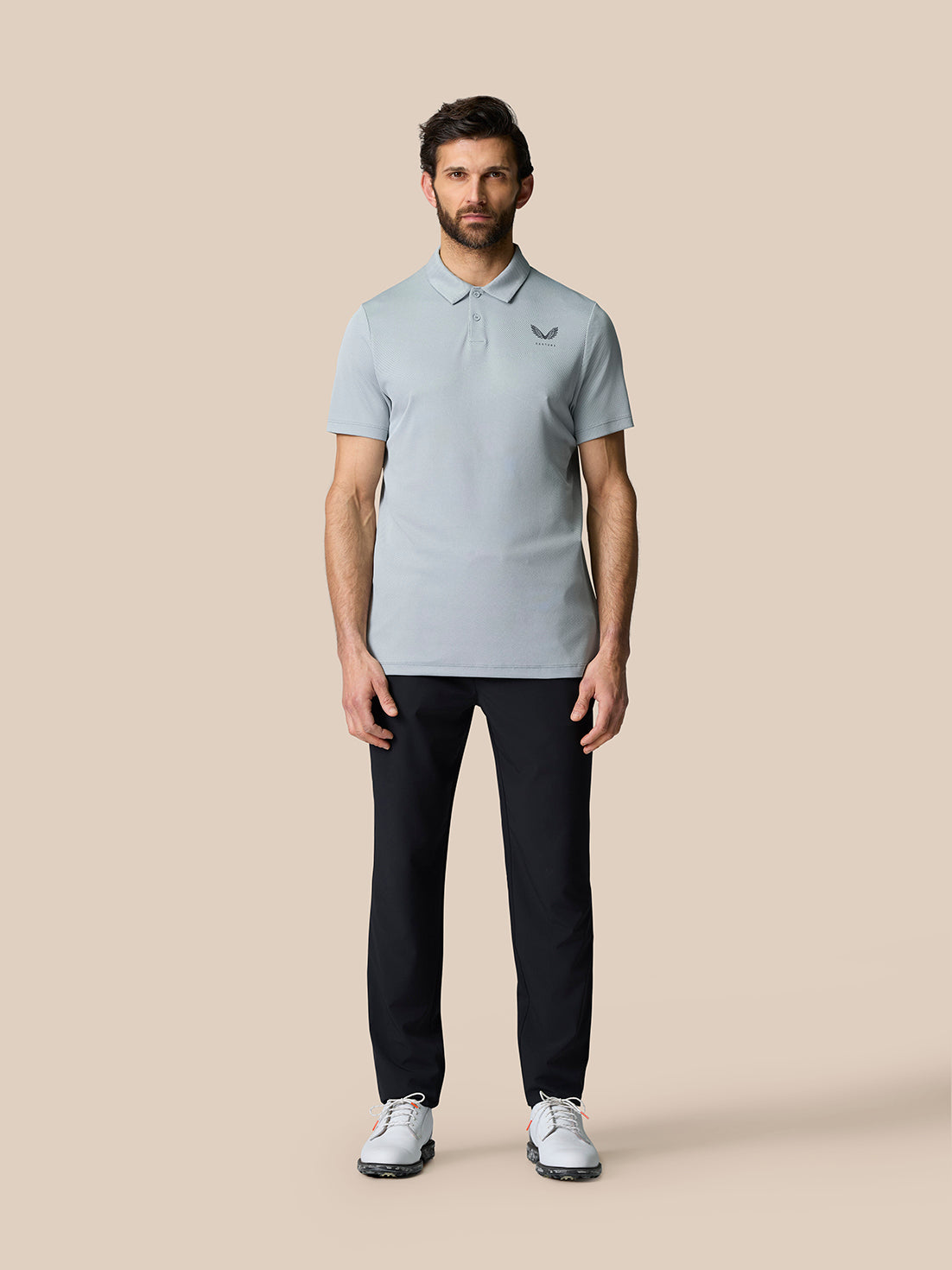 Full body shot of a man in a light grey polo top with CASTORE logo on chest and dark trousers, standing in neutral pose