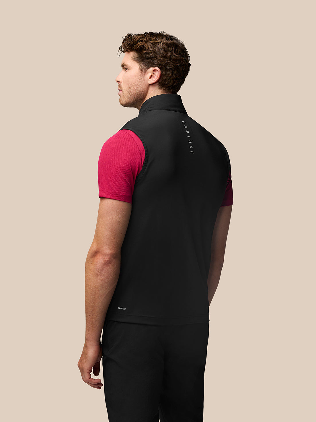 Male model wearing black Golf Pinnacle Gilet over a pink short-sleeve top; stand collar, sleeveless, with Castore branding down the back and Protek at the lower back.