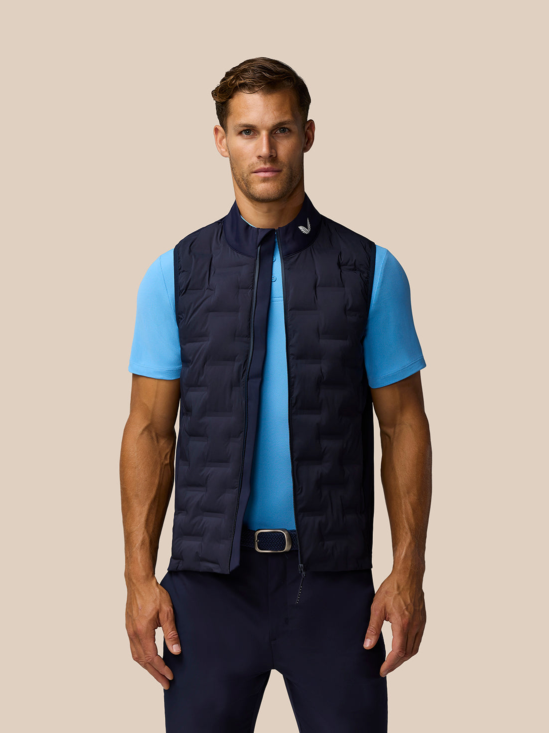 Male model wearing navy Pinnacle Gilet over a light blue polo against a beige studio backdrop