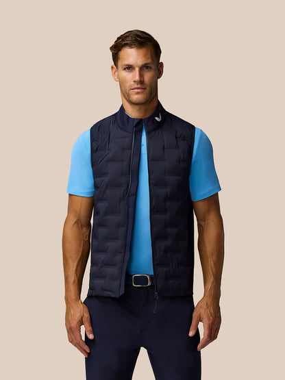 Male model wearing navy Pinnacle Gilet over a light blue polo against a beige studio backdrop