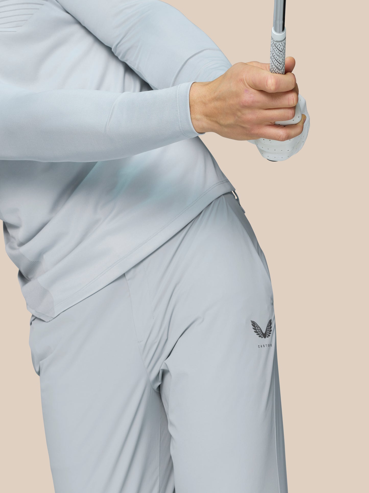 Side view of a person wearing light grey Golf Pinnacle Trousers with leg zip closures and Castore logo on the thigh