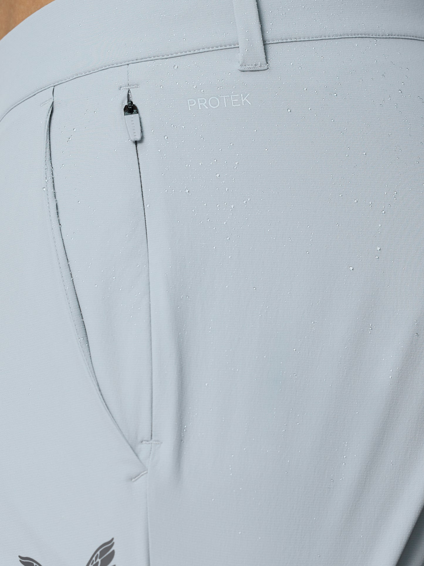 Close up of light grey golf trousers fabric with water droplets, PROTEK branding, belt loop and small zip pocket.