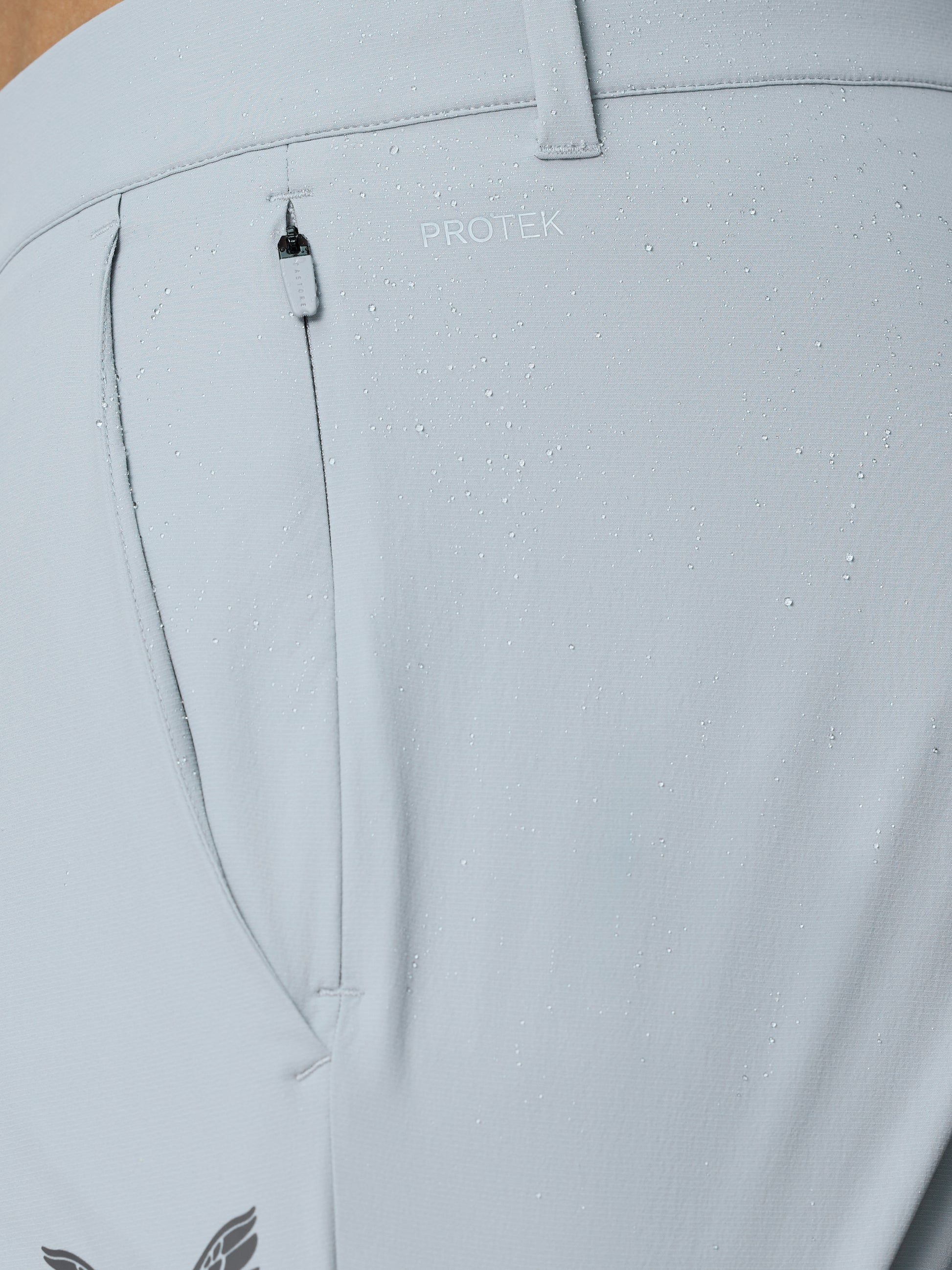 Close up of light grey golf trousers fabric with water droplets, PROTEK branding, belt loop and small zip pocket.