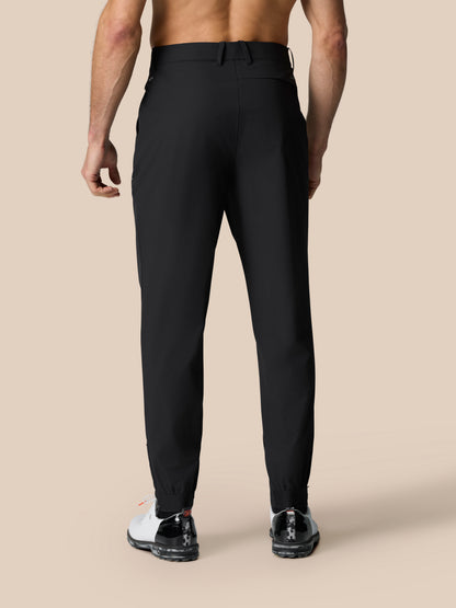 Back view of a man wearing black Golf Pinnacle Trousers with belt loops and elastic cuffs against a beige backdrop.
