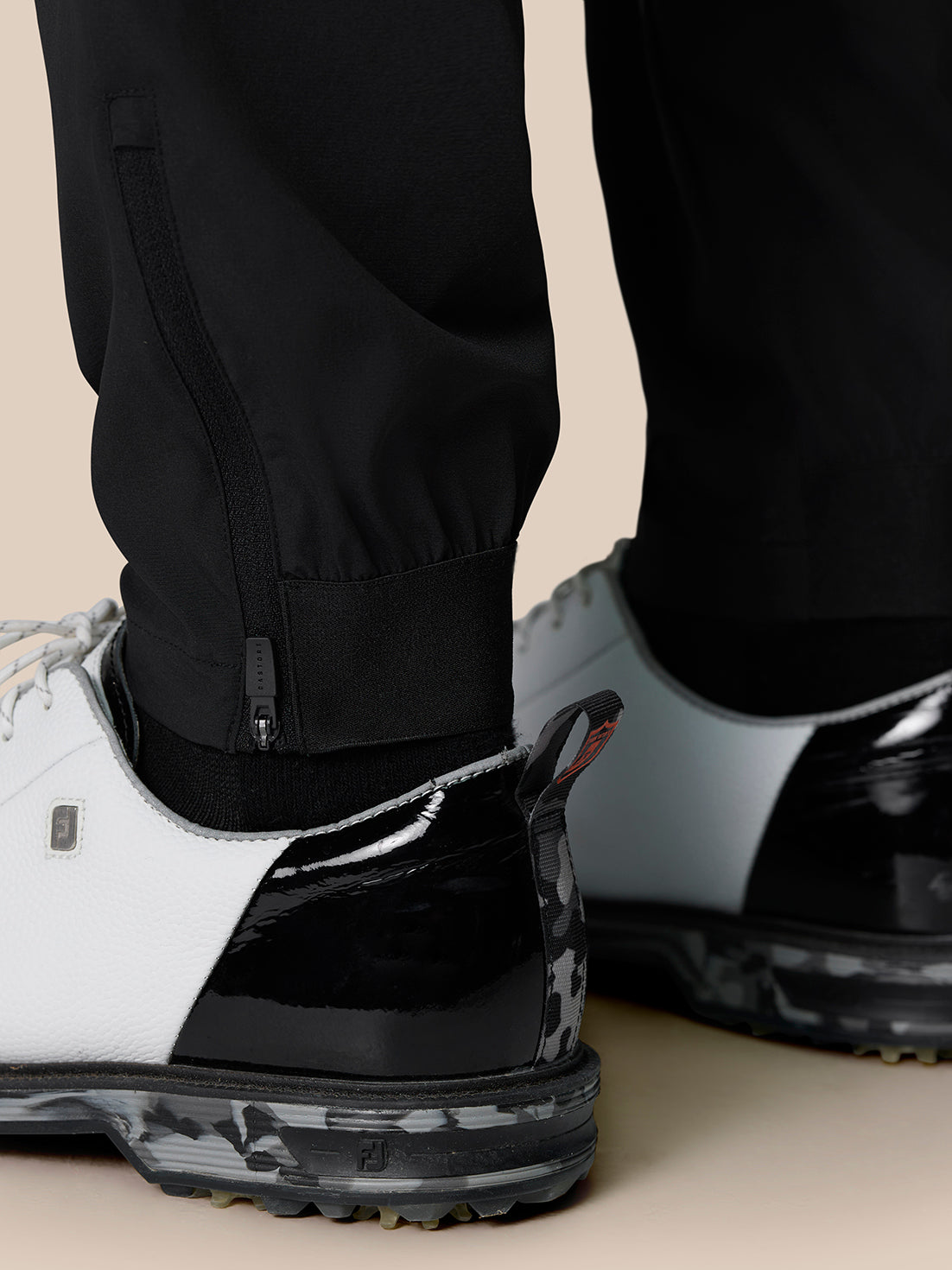 Close-up of the lower leg with ankle zip closures and elastic cuff on black Golf Pinnacle Trousers worn with white and black sneakers.