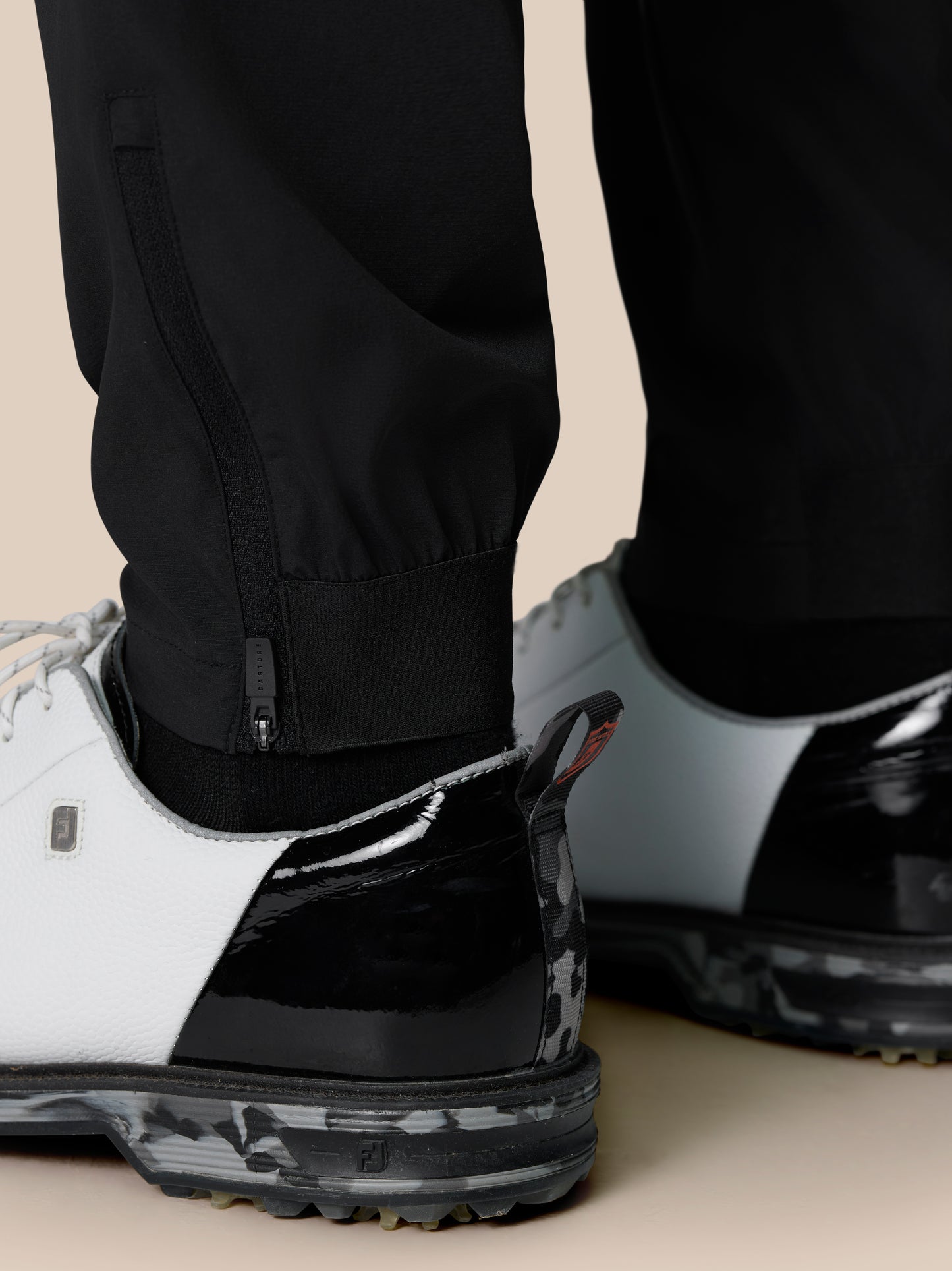 Close up of the leg zipper area on black Golf Pinnacle Trousers with Castore branding near the ankle and a white sneaker.