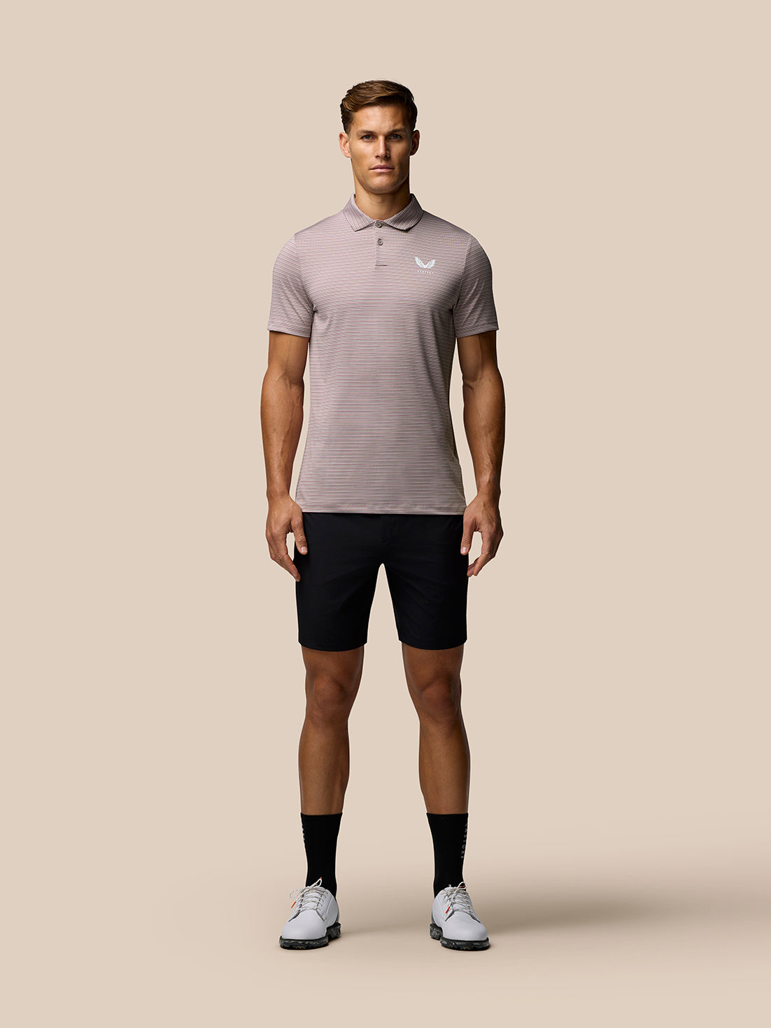 Full-body front view of a male model wearing pink yarn-dye stripe polo with white Castore logo, black shorts and white sneakers.