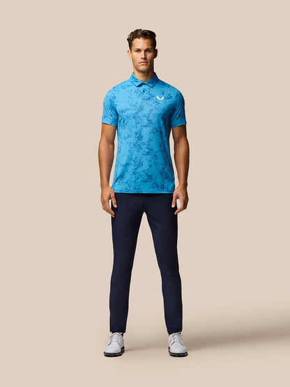 Male model in blue geo print golf polo showing Castore branding on chest against beige backdrop