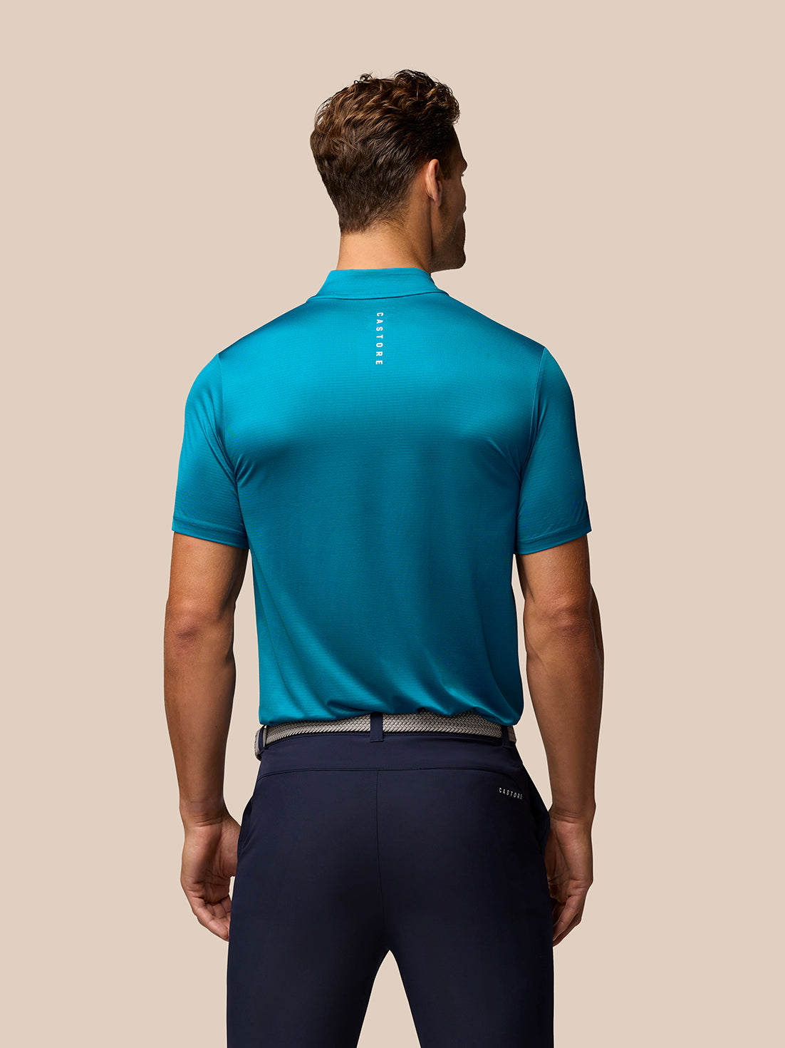 Back view of a male model wearing a teal Pinnacle Golf Varsity Polo with vertical Castore branding down the back