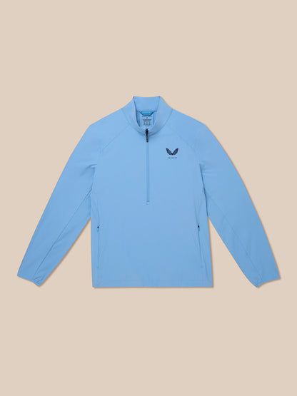 Flat lay of a sky blue half zip golf jacket with Castore logo on the chest and zip pockets on a beige background