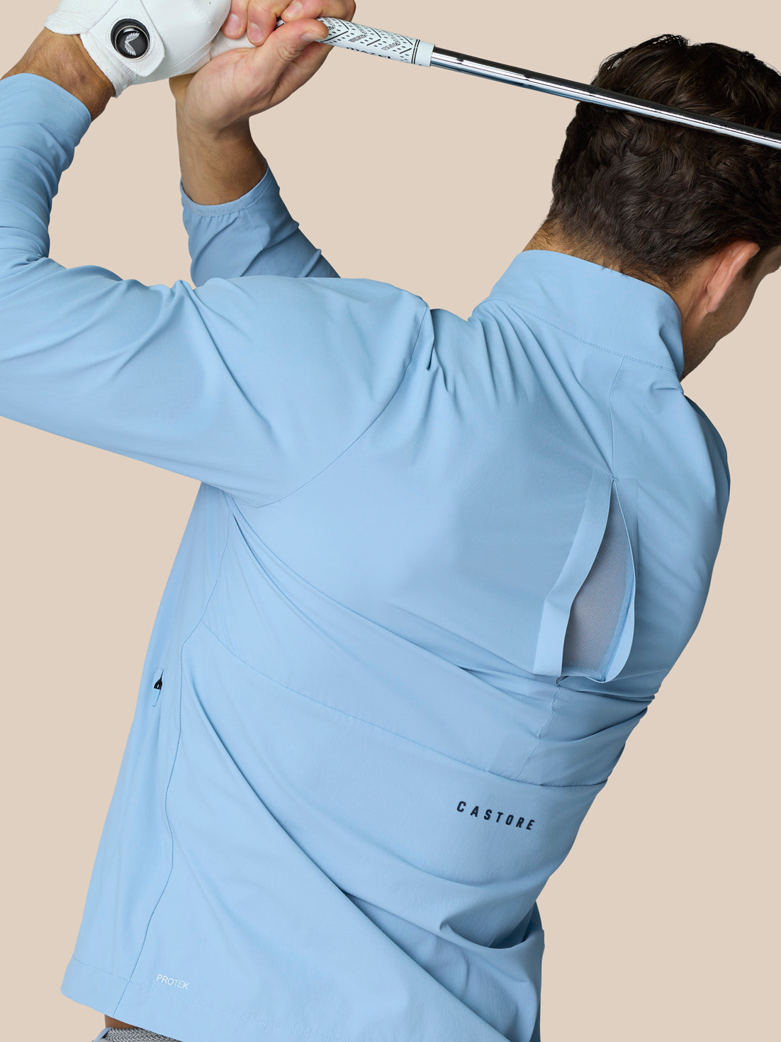 Back view of a man in a sky blue golf jacket mid swing with a golf club; Castore logo on the lower back