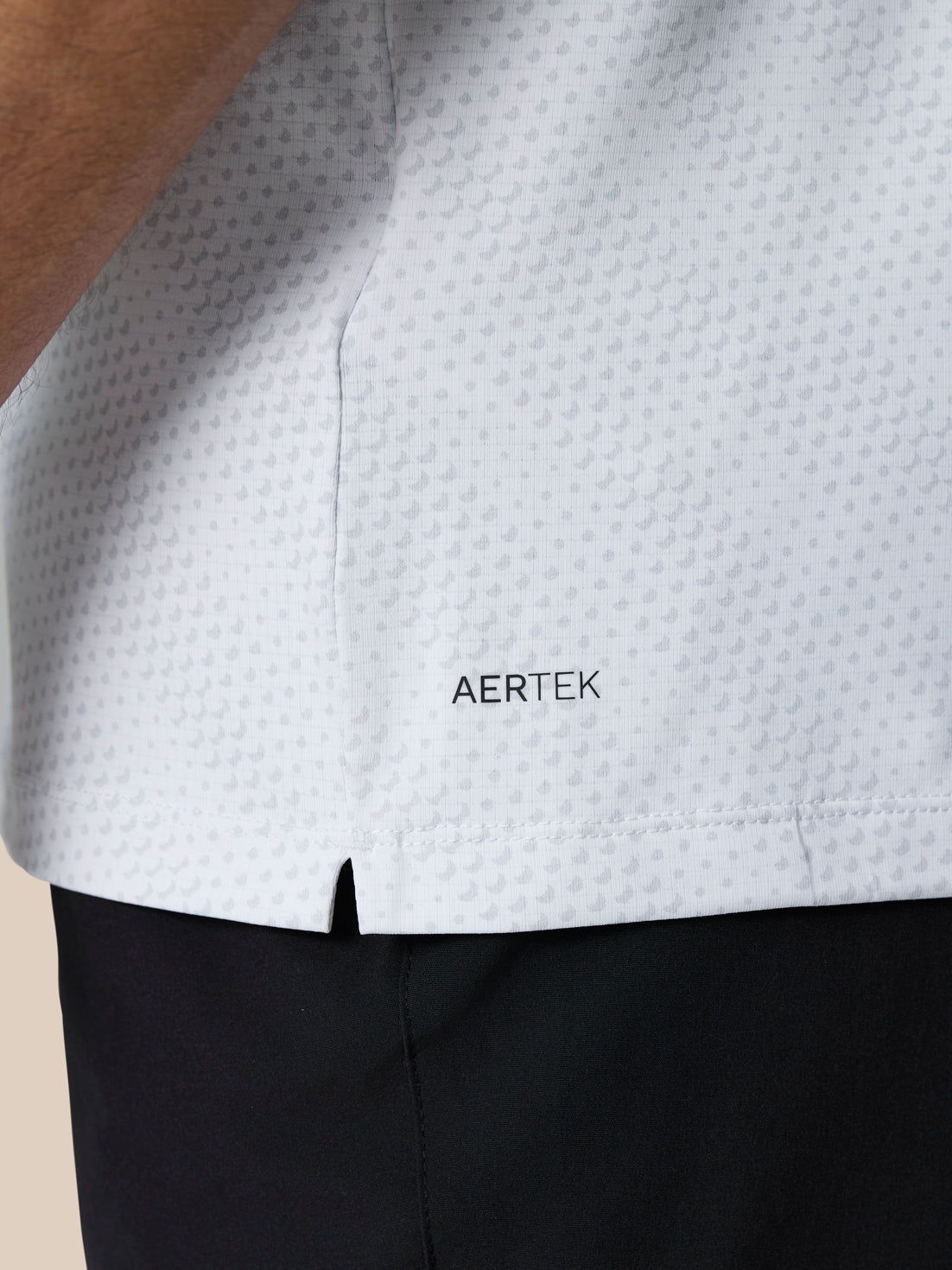 Close up of the lower back hem of the white Club Graphic Polo showing AERTEK branding and a small side vent slit.