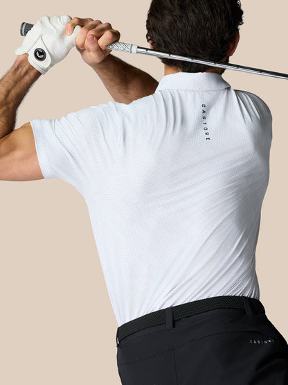 Back view of a male golfer in a white dotted polo with Castore branding, gripping a golf club across his shoulders.