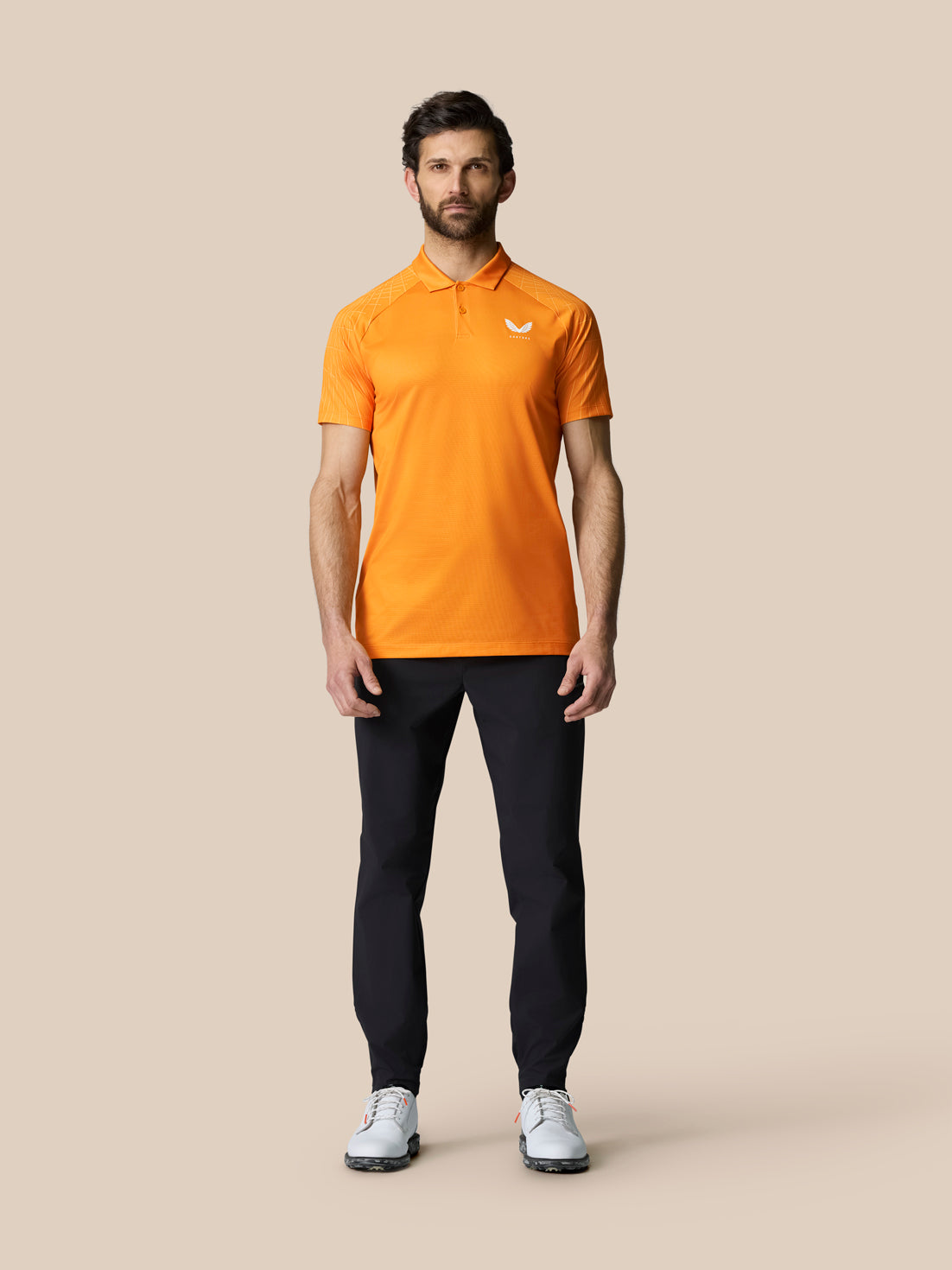 Front view of a man in an orange Club Graphic Polo with sleeve graphic; Castore logo on chest, black trousers and white golf shoes.