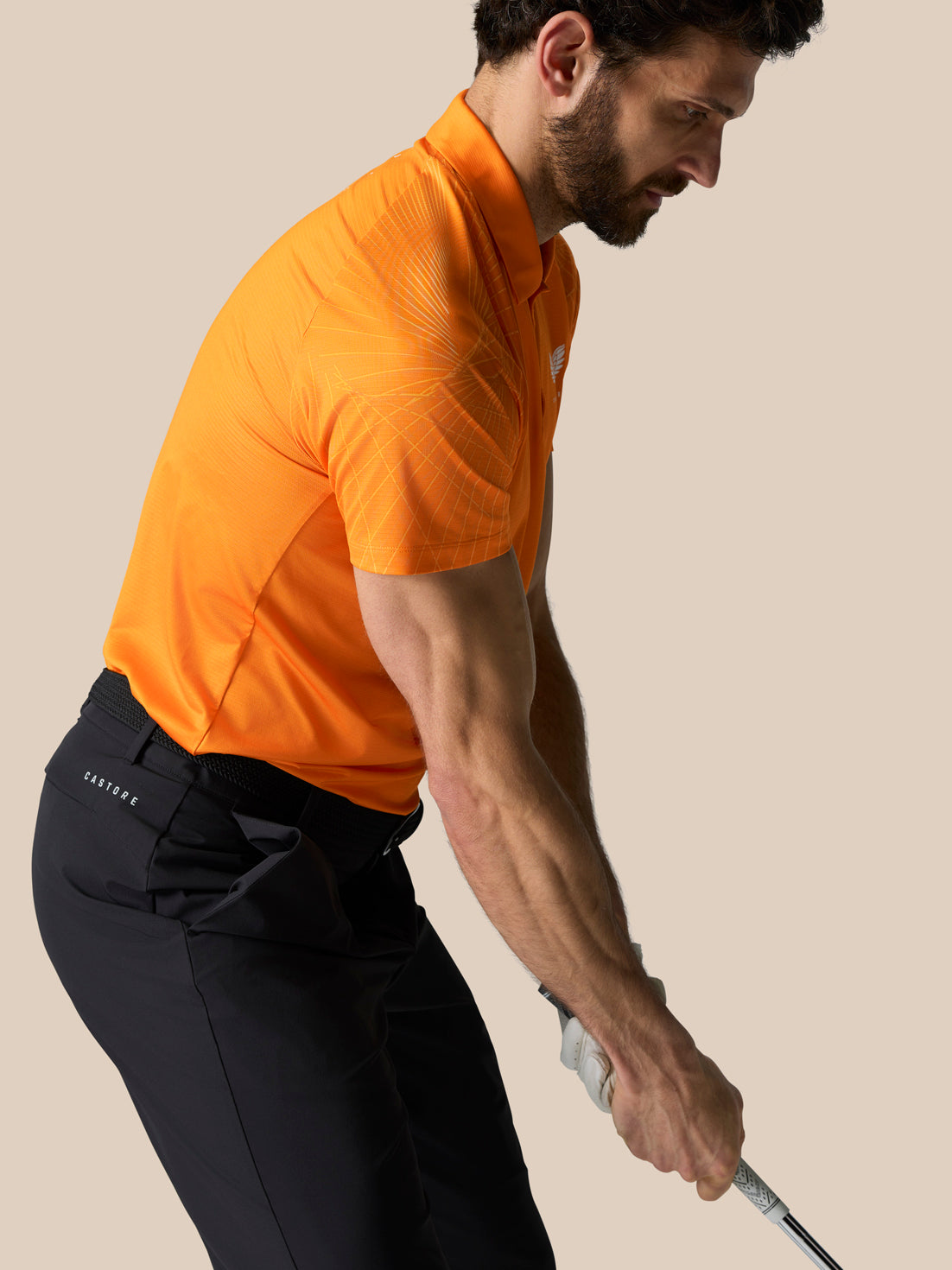 Golfer wearing bright orange Club Graphic Polo Mango bending forward and gripping a golf club; sleeve graphic and Castore logo on chest.