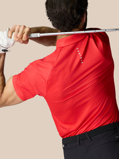 Back view of a golfer wearing a red Castore Club Graphic Polo with branding on the chest and upper back, holding a golf club across the shoulders.