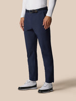 Close-up of navy Golf Pinnacle trousers at the waist with a black belt against a beige background.