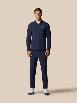 Male model wearing a navy golf outfit featuring a zip front jacket and matching trousers against a beige backdrop.
