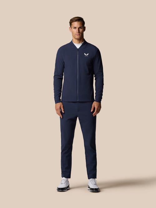 Male model wearing a navy golf outfit featuring a zip front jacket and matching trousers against a beige backdrop.
