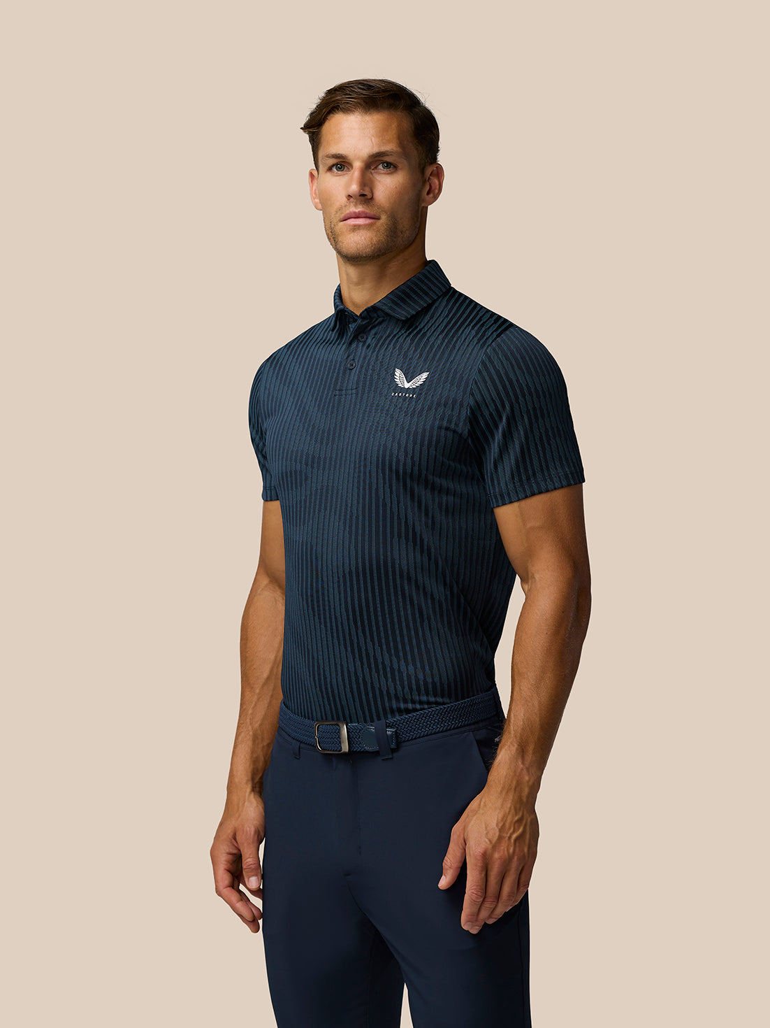 Three quarter view of male model wearing navy striped Golf Pinnacle Glow Polo with navy trousers and belt