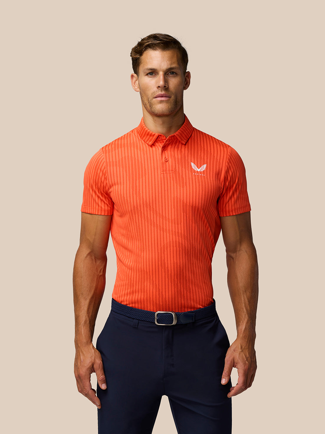 Male model wearing bright orange striped golf polo with Castore logo on left chest and navy pants