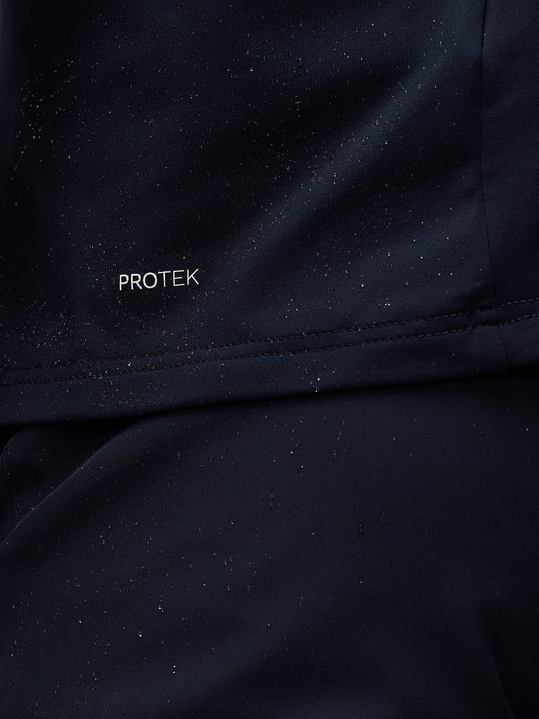 Close-up of navy jacket fabric showing PROTEK branding with water droplets on the surface