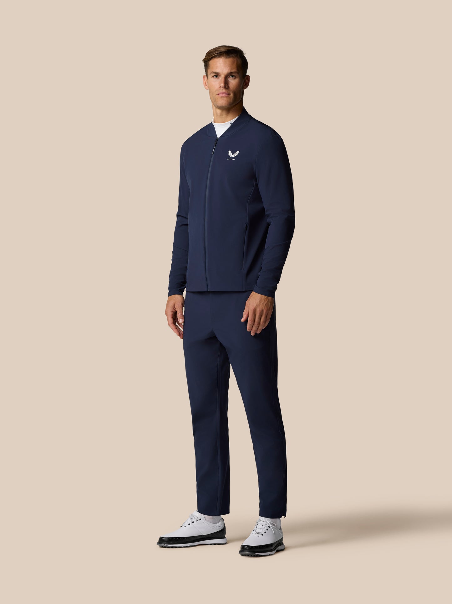 Full-length shot of a man in the navy Golf Pinnacle LVS Jacket with white sneakers and a belt.