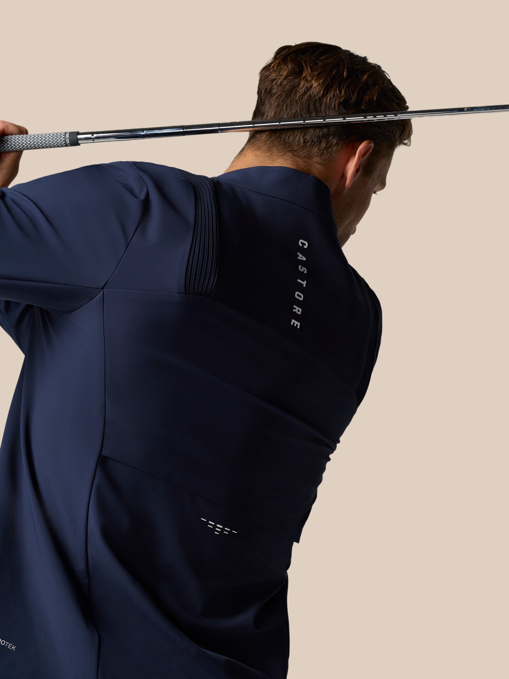 Man wearing midnight navy Castore Layered Ventilation System jacket during golf backswing with a club against a beige background